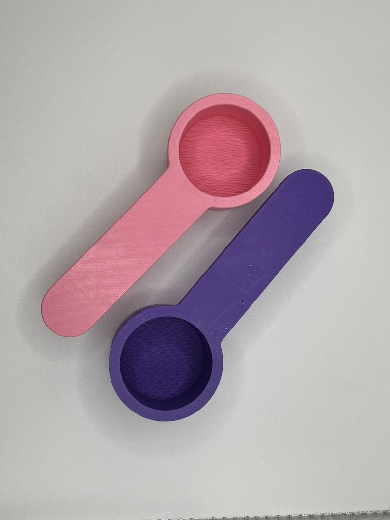Peut inclure: Deux cuill&egrave;res doseuses avec des bols ronds et des manches plats. Une cuill&egrave;re est rose clair et l'autre est violet fonc&eacute;. Les deux articles pr&eacute;sentent une finition de surface textur&eacute;e typique des objets imprim&eacute;s en 3D, avec un bol circulaire.