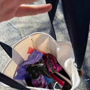Pode incluir: Uma sacola de lona com al&ccedil;as pretas, cheia de produtos de higiene. A sacola cont&eacute;m um borrifador, escovas, pentes e embalagens. O texto "What's Inside" &eacute; vis&iacute;vel em uma embalagem roxa.