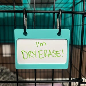 May include: A turquoise dry erase board with a white surface and black clips hanging on a black metal cage. The board says "I'm Dry Erase!" in green marker.
