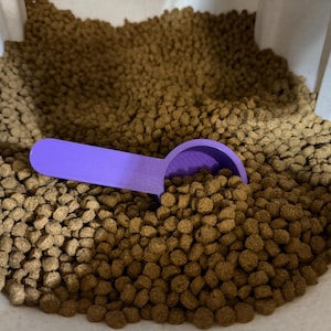 Peut inclure: Une pelle en plastique violet vif repose dans un r&eacute;cipient rempli de petites croquettes rondes et brunes pour animaux. La pelle pr&eacute;sente un long manche et un bol circulaire profond, partiellement immerg&eacute; dans le tas de morceaux de nourriture.