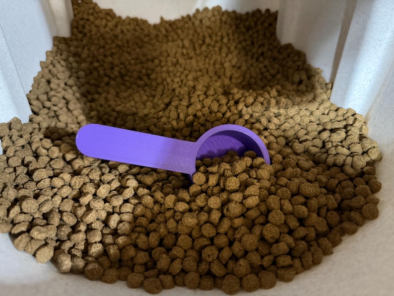 Peut inclure: Une pelle en plastique violet vif repose dans un r&eacute;cipient rempli de petites croquettes rondes et brunes pour animaux. La pelle pr&eacute;sente un long manche et un bol circulaire profond, partiellement immerg&eacute; dans le tas de morceaux de nourriture.