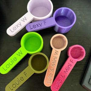 May include: Six colorful measuring spoons with personalized names. The spoons are made of plastic and have a scoop shape. The names on the spoons are "Lexy W", "Lexy F", "Louie W", "Louie F", "Lula & Mini W", and "Lula & Mini F".