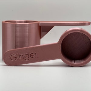 May include: A rose gold colored measuring spoon with a handle that says "Ginger" on it.