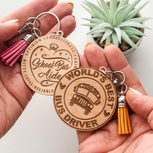 May include: Two wooden keychains with a school bus theme. One keychain is heart-shaped and says "School Bus Aide" with a small school bus image. The other keychain is round and says "World's Best Bus Driver" with a larger school bus image. Both keychains have a small tassel.
