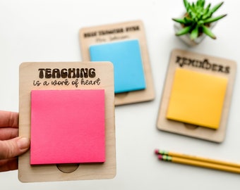 May include: A wooden note holder with the text "TEACHING is a work of heart" engraved on it. A pink sticky note is attached to the holder. The holder is on a white surface.
