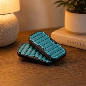 Puede incluir: Dos objetos rectangulares de color verde azulado y negro con una superficie acanalada están apilados sobre una mesa de madera. Los objetos tienen un borde negro y un interior verde azulado con líneas paralelas. Los objetos son probablemente un par de accesorios.