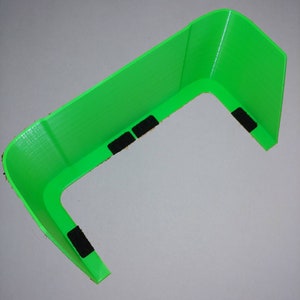 May include: Bright green plastic visor with a curved design. The visor has a rectangular shape with rounded corners and several black velcro strips attached to the underside. The visor is designed to provide shade and protection from the sun.