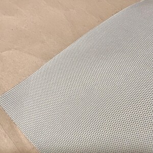May include: A close-up shot of a light gray woven fabric with a textured, grid-like pattern. The fabric is angled across the frame, with a tan surface in the background. The fabric appears to be a durable material.