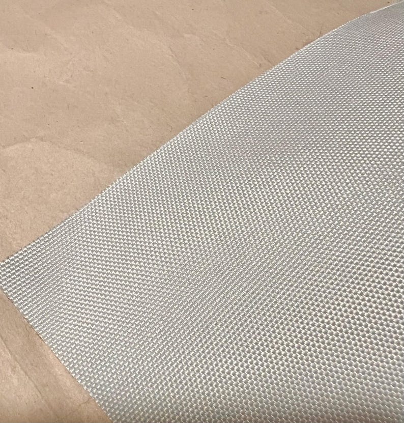 May include: A close-up shot of a light gray woven fabric with a textured, grid-like pattern. The fabric is angled across the frame, with a tan surface in the background. The fabric appears to be a durable material.