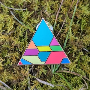 Puede incluir: Un pin triangular de esmalte con un patrón geométrico de formas segmentadas en colores vibrantes que incluyen cian, magenta, verde lima, naranja, azul oscuro y rosa, todo delineado con finas líneas metálicas sobre una superficie plana.