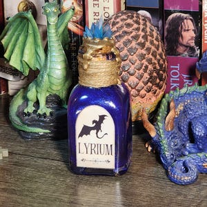 May include: A dark blue, glittery potion bottle with a label that reads "LYRIUM" and a dragon illustration. The bottle is adorned with a gold-colored stopper and twine. A green dragon figurine and fantasy books are in the background.