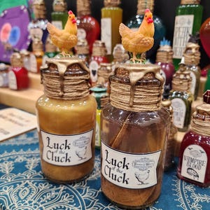 May include: Two glass bottles with cork stoppers, twine, and gold wax seals. Each bottle has a small chicken figurine on top. The labels read "Luck of the Cluck" with a chicken illustration. The bottles are surrounded by other decorative bottles.