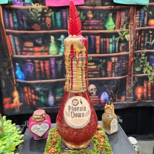 May include: A decorative bottle labeled "Phoenix Down" with a red and gold design. The bottle has a red feather on top and is surrounded by other potion bottles. The background features a bookshelf.