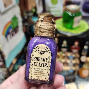 May include: A small, purple glass bottle filled with glitter and a cork stopper. The label reads "Sneaky Elixir" with text below that says "Allows you to move about undetected by monsters and animals." The bottle is sealed with a gold-colored wax and twine.