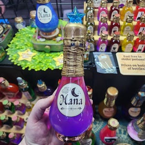 May include: A decorative potion bottle with a purple and pink liquid, a gold-colored neck, and a blue star-shaped stopper. The bottle has a label that says "Mana" with a crescent moon design. Other bottles are visible in the background.