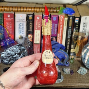 May include: A red glass bottle with a glittery interior, labeled "Phoenix Down." The bottle has a gold-colored twine neck and a red, pink, and gold stopper. The background includes a shelf of books.