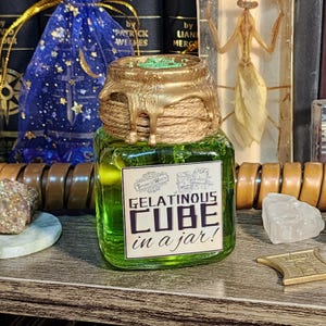 May include: A square glass jar filled with a vibrant green liquid, labeled "GELATINOUS CUBE in a jar!". The jar has a gold-colored, dripped wax seal and a twine-wrapped lid. The background includes books and a blue drawstring bag.