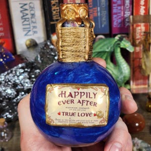 May include: A blue potion bottle with a gold-colored top and a label that reads "Happily Ever After" and "True Love." The bottle has a textured, metallic appearance. The top is adorned with gold-colored embellishments and wrapped with twine.