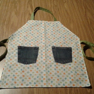 May include: A white apron with a green trim and two denim pockets. The apron has a white background with a small, colorful pattern of leaves and dots.