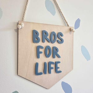 May include: Wooden wall hanging with the words "BROS FOR LIFE" in blue, 3D-style letters. The sign is a light tan color and is suspended by a natural rope. The background is white with blue and tan paint splotches.