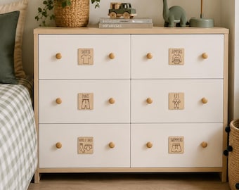 May include: A light wood and white six-drawer dresser featuring wooden knobs and square labels on each drawer. The labels display text and icons for SHIRTS, JUMPERS, PANTS, PJ'S, JOCKS & SOCKS, and SWIMMERS. The dresser has a clean, rectangular frame.