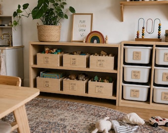 May include: A light wood open shelving unit containing six wooden storage crates labeled with text and icons for blocks, trains, wood toys, books, soft toys, and small world. Beside it, a white storage unit holds white bins with labels.