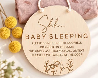 May include: A round wooden sign with a white rope hanger featuring the engraved text Shh... BABY SLEEPING, PLEASE DO NOT RING THE DOORBELL OR KNOCK ON THE DOOR, WE KINDLY ASK THAT YOU CALL OR TEXT, PLEASE LEAVE PARCELS AT THE DOOR, and floral art.