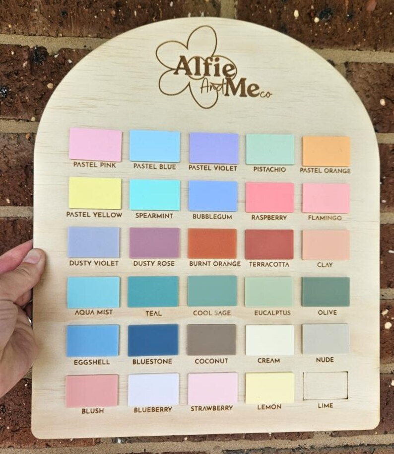 May include: Wooden color palette with 25 pastel color swatches, including pink, blue, violet, and orange. The top of the palette has the text "Alfie And Me co" in a floral design. The color names are printed below each swatch.