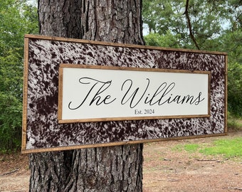 Op de afbeelding: Een houten bord met een bruin en wit koehuidpatroonframe. Het bord heeft een witte achtergrond met zwarte tekst die "The Williams Est. 2024" luidt.