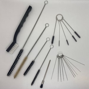May include: A selection of cleaning brushes and tools. Includes a black-handled brush with white bristles, several long, thin brushes with looped ends, and a set of small brushes attached to a ring. The brushes have black, beige, and silver bristles.
