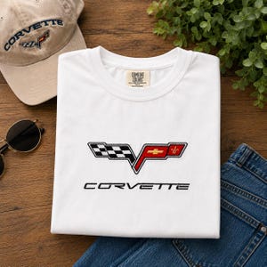 Puede incluir: Una camiseta blanca de cuello redondo con el logotipo de Corvette bordado en el pecho. El logo incluye una bandera a cuadros blanca y negra y una sección roja con un emblema dorado de Chevrolet y una flor de lis. La palabra Corvette está bordada debajo.