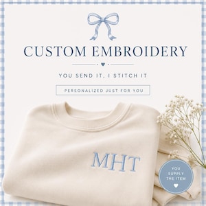 Op de afbeelding: Een crèmekleurig sweatshirt met lichtblauw geborduurde initialen MHT op de borst. De tekst luidt CUSTOM EMBROIDERY, YOU SEND IT, I STITCH IT, PERSONALIZED JUST FOR YOU en YOU SUPPLY THE ITEM. Een lichtblauw striksymbool staat bovenaan.