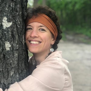 May include: A solid rust-colored fabric headband worn across the forehead, paired with dangling earrings featuring circular white and dark gray components. The headband is made of a smooth, stretchy material and sits securely above the ears.