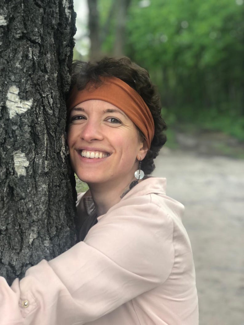 May include: A solid rust-colored fabric headband worn across the forehead, paired with dangling earrings featuring circular white and dark gray components. The headband is made of a smooth, stretchy material and sits securely above the ears.