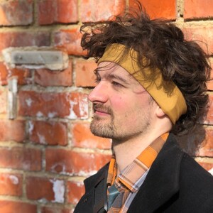 May include: A mustard yellow fabric headband worn around the head, featuring a smooth, solid-colored texture. The headband is positioned across the forehead, resting against dark, curly hair, and is made of a flexible, stretchable material.