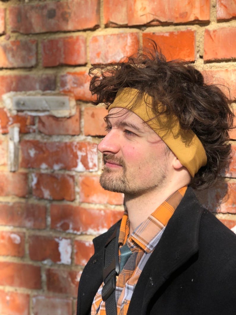 May include: A mustard yellow fabric headband worn around the head, featuring a smooth, solid-colored texture. The headband is positioned across the forehead, resting against dark, curly hair, and is made of a flexible, stretchable material.