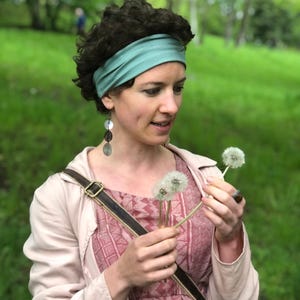 May include: A sage green fabric headband, a light pink jacket, and a pink patterned dress. The outfit includes long dangling earrings with circular discs and a brown leather belt with a metal buckle. The attire is worn with a brown leather bag strap.
