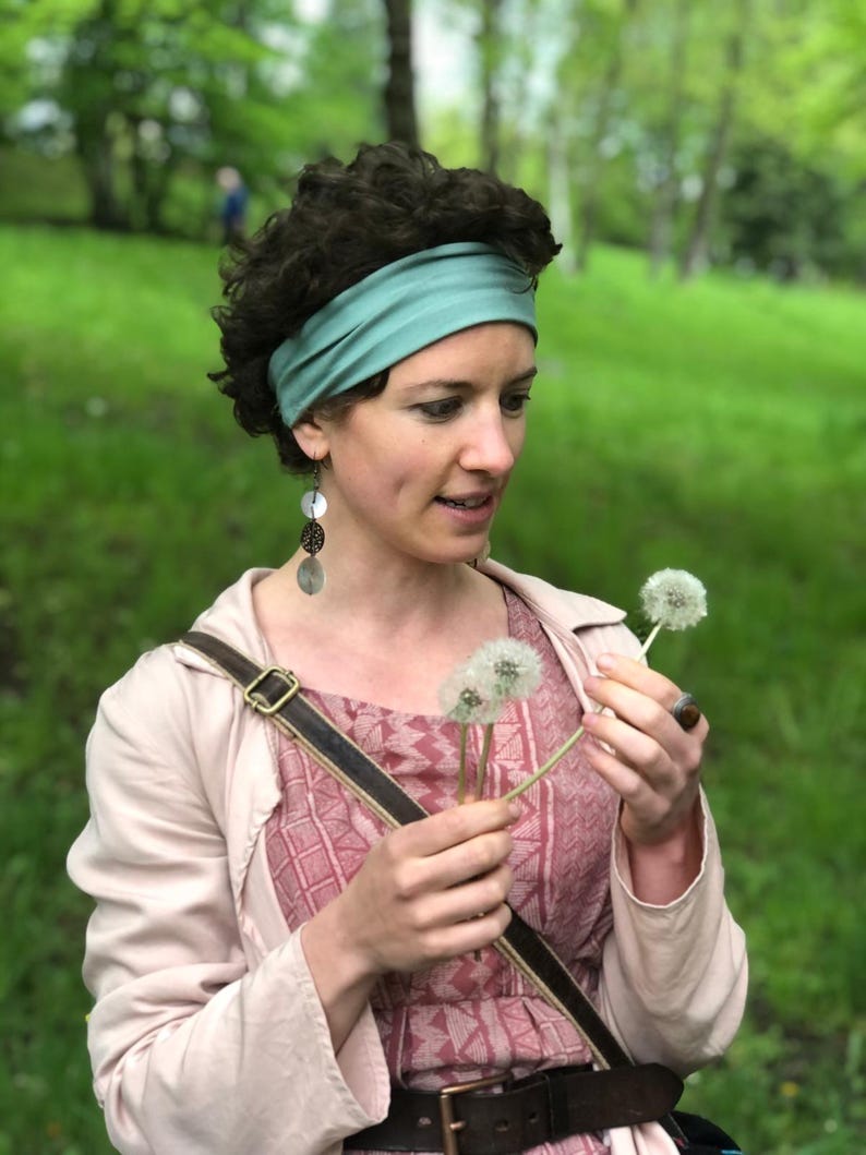 May include: A sage green fabric headband, a light pink jacket, and a pink patterned dress. The outfit includes long dangling earrings with circular discs and a brown leather belt with a metal buckle. The attire is worn with a brown leather bag strap.