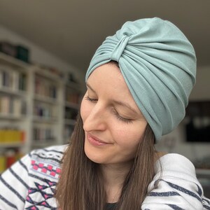 May include: A sage green turban-style head covering made of soft, stretchy fabric. The headpiece features a gathered, pleated design at the front center, creating a structured, layered look that fits snugly around the head with a smooth finish.
