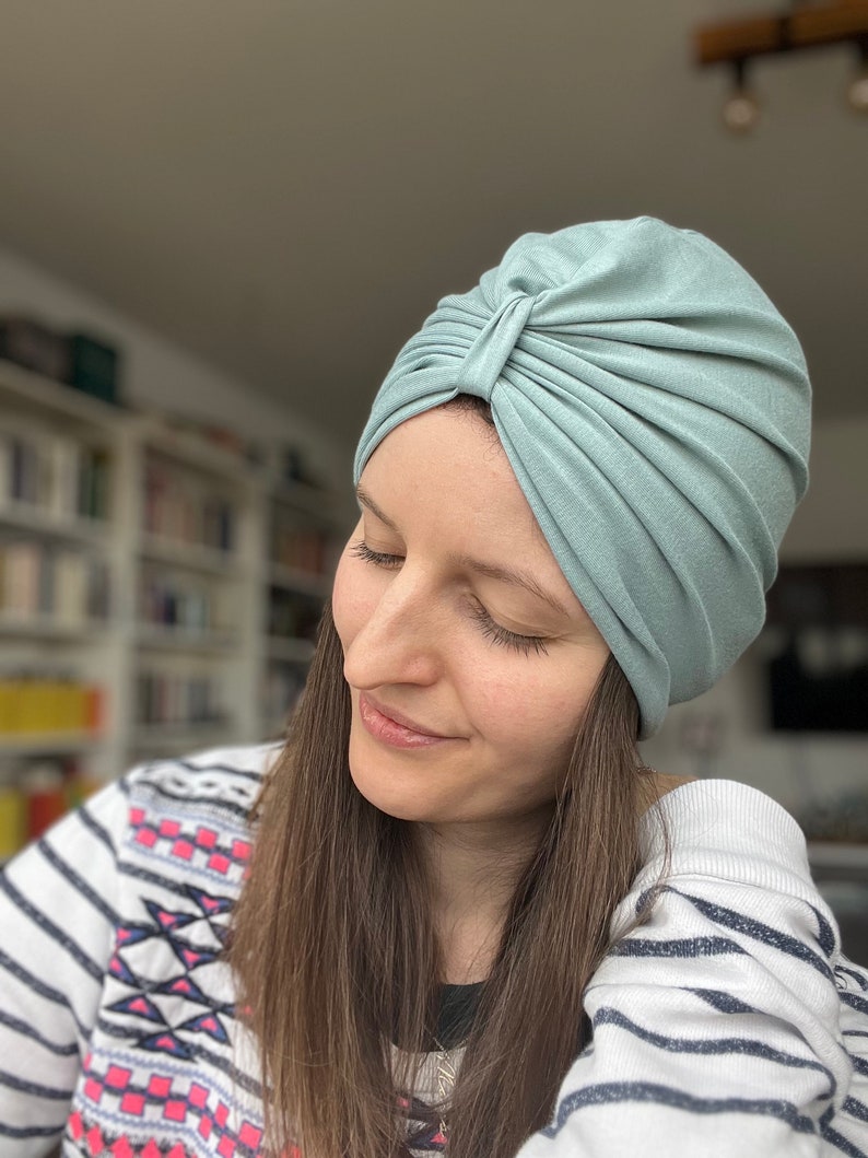 May include: A sage green turban-style head covering made of soft, stretchy fabric. The headpiece features a gathered, pleated design at the front center, creating a structured, layered look that fits snugly around the head with a smooth finish.