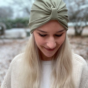 May include: A sage green turban-style head covering made of soft fabric. The cap features a gathered, pleated design at the front center, creating a knotted appearance with radiating folds that extend across the crown for a fitted look.