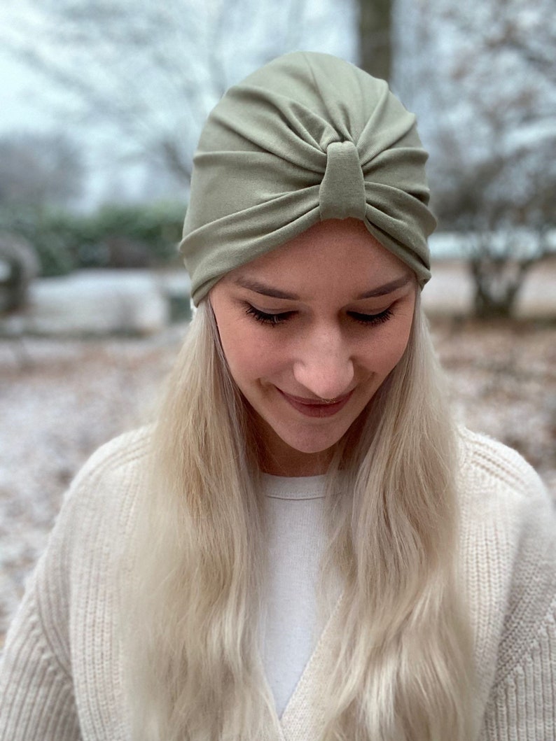 May include: A sage green turban-style head covering made of soft fabric. The cap features a gathered, pleated design at the front center, creating a knotted appearance with radiating folds that extend across the crown for a fitted look.
