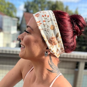 May include: A wide fabric headband featuring a cream-colored base with a repeating pattern of orange and brown roses, leaves, and delicate botanical sprigs. The headband is worn around the head, covering the forehead and ears with a soft, smooth texture.