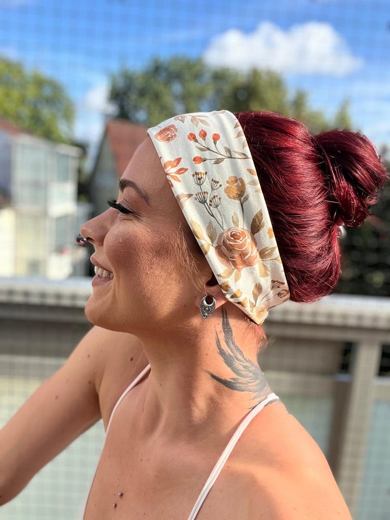 May include: A wide fabric headband featuring a cream-colored base with a repeating pattern of orange and brown roses, leaves, and delicate botanical sprigs. The headband is worn around the head, covering the forehead and ears with a soft, smooth texture.
