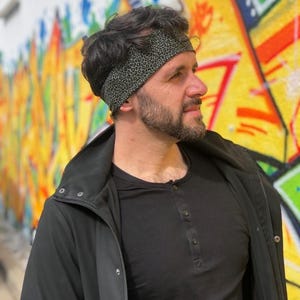 May include: A wide headband featuring a dark olive green fabric with a small black leopard print pattern. The headband is worn around the head, resting across the forehead and over the hair, providing a snug fit against the scalp.