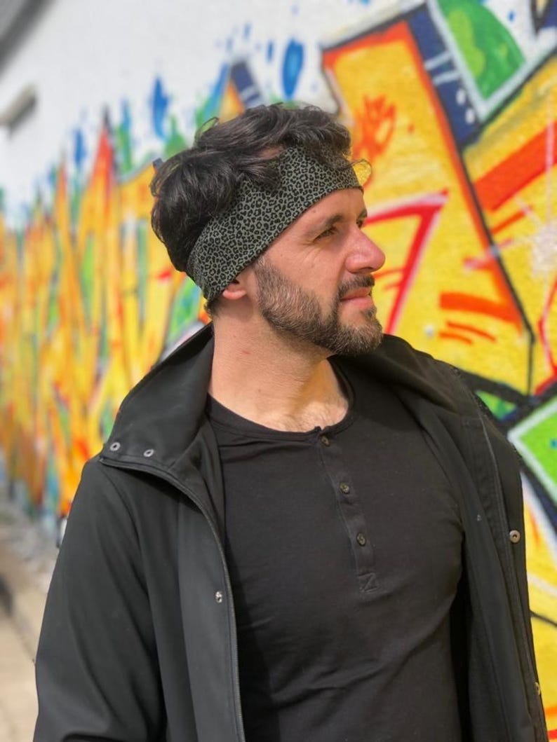 May include: A wide headband featuring a dark olive green fabric with a small black leopard print pattern. The headband is worn around the head, resting across the forehead and over the hair, providing a snug fit against the scalp.