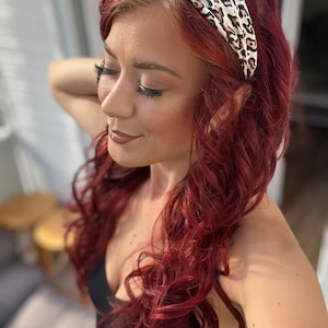 May include: A wide fabric headband featuring a cream-colored base with a brown and black leopard print pattern. The headband is worn across the top of the head, resting against long, wavy, deep red hair that falls over the shoulders.