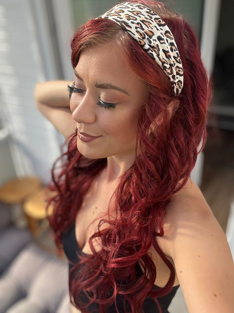 May include: A wide fabric headband featuring a cream-colored base with a brown and black leopard print pattern. The headband is worn across the top of the head, resting against long, wavy, deep red hair that falls over the shoulders.