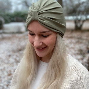 May include: A sage green turban-style hat featuring a gathered front detail with a central knot. The headwear is constructed from a smooth, solid-colored fabric that fits snugly around the head, showcasing soft pleats radiating from the center.
