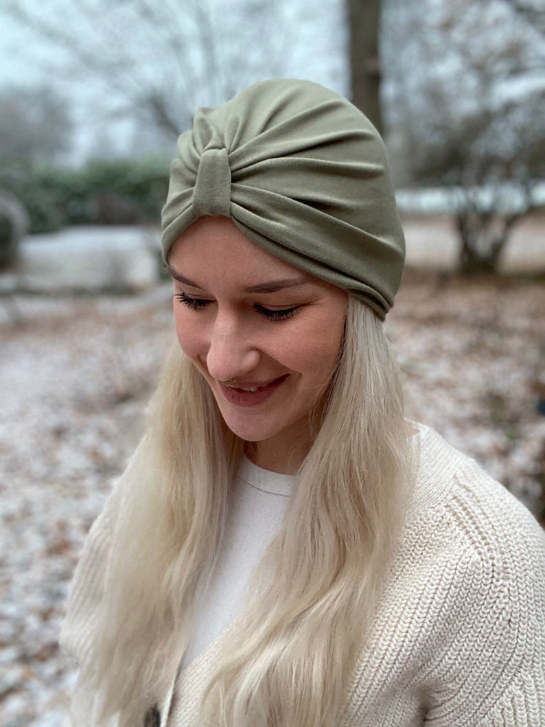 May include: A sage green turban-style hat featuring a gathered front detail with a central knot. The headwear is constructed from a smooth, solid-colored fabric that fits snugly around the head, showcasing soft pleats radiating from the center.